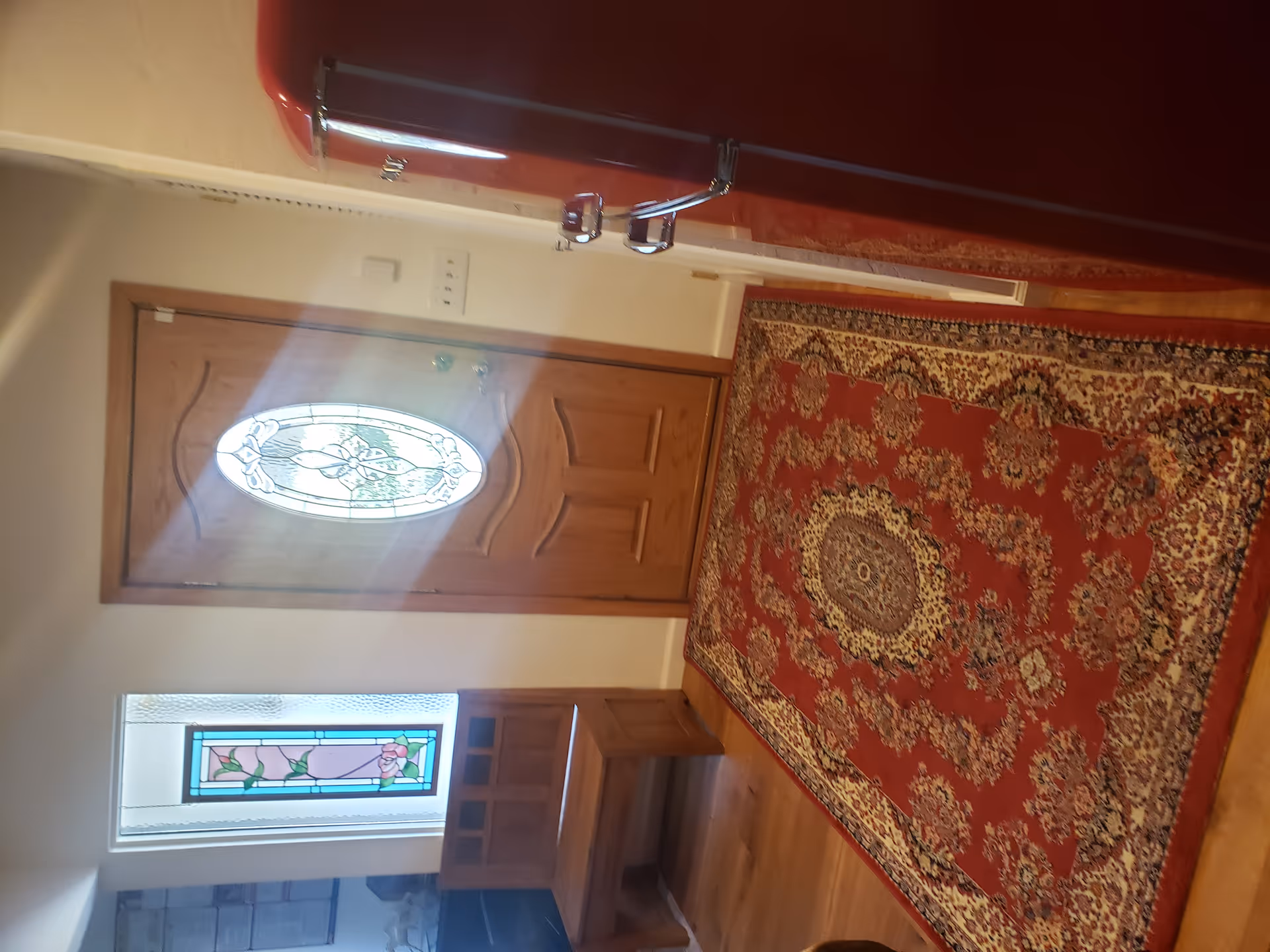 Interior view of an entryway featuring a wooden door with an oval decorative glass panel. To the left of the door is a stained glass window with floral designs and a wooden bench beneath it. A red patterned rug covers the wooden floor, and a red retro-style refrigerator is visible on the right side.