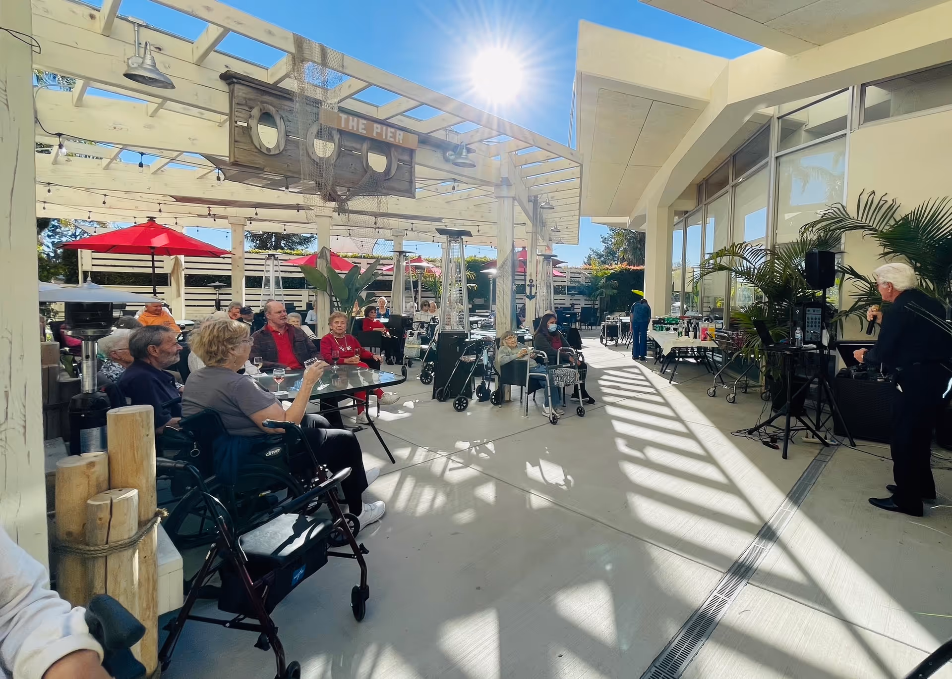 Outdoor patio area at Ventura Townehouse with elderly residents seated at tables under a pergola with red umbrellas. A man is speaking into a microphone near a sound system setup. The sun shines brightly in a clear blue sky, casting shadows on the ground.