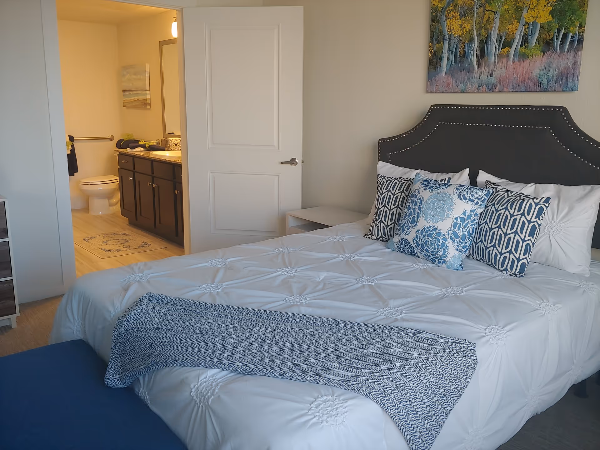 A bedroom with a neatly made bed featuring a dark upholstered headboard, white bedding, and decorative blue and white pillows. A blue throw blanket is draped across the foot of the bed. To the left, an open door reveals a bathroom with a toilet, dark wood vanity, and a mirror. A colorful nature painting hangs on the wall above the bed.