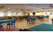 Spacious dining room with multiple tables covered in green tablecloths and chairs arranged around them. The room has wooden flooring, ceiling fans, and bright lighting with windows along the walls.