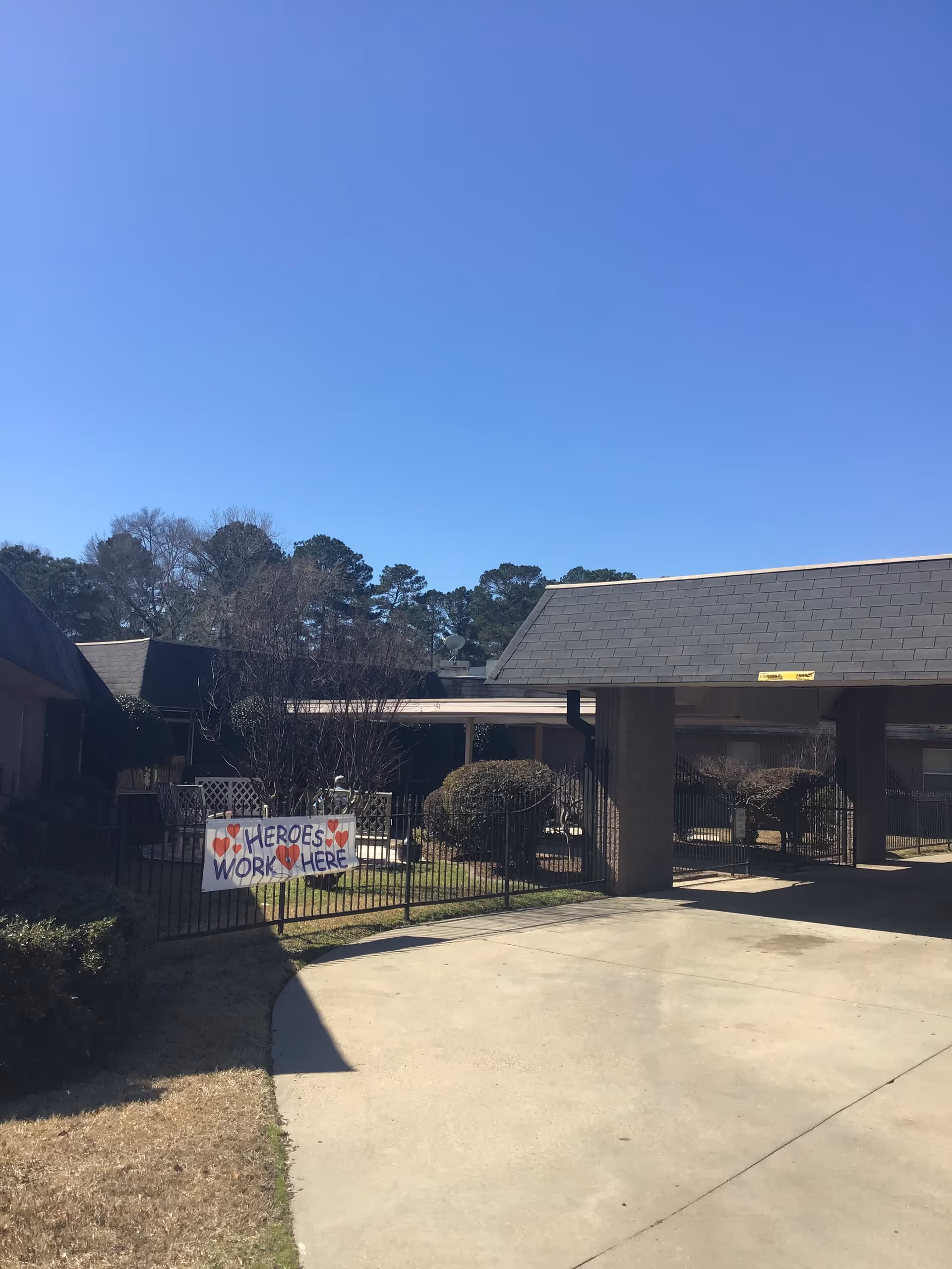 Exterior front entrance of a nursing facility showing a driveway, fenced courtyard and a handmade "Heroes Work Here" sign.