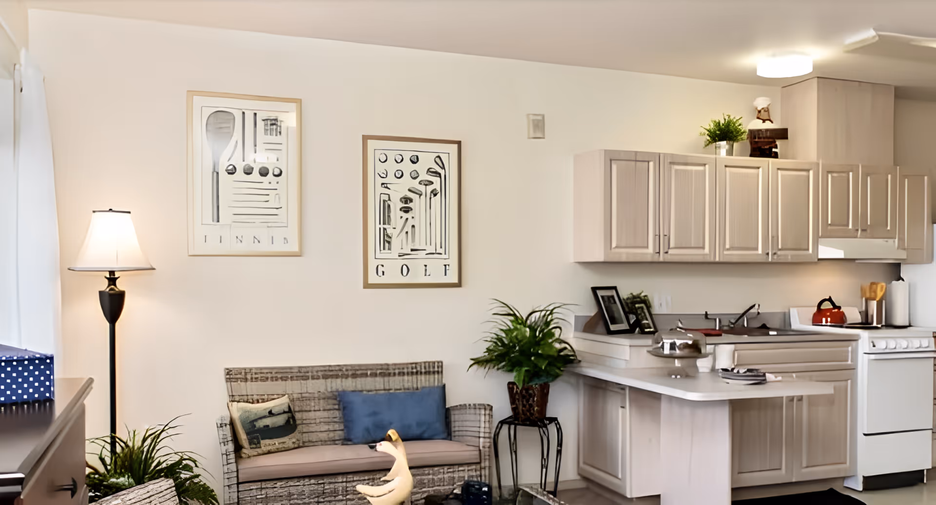 A cozy living area with a small couch adorned with two pillows, a floor lamp, and two framed artworks on the wall labeled 'TENNIS' and 'GOLF'. Adjacent to the living area is a compact kitchen with light-colored cabinets, a countertop with decorative items, a stove, and a red kettle. There are also some green plants adding a touch of nature to the space.