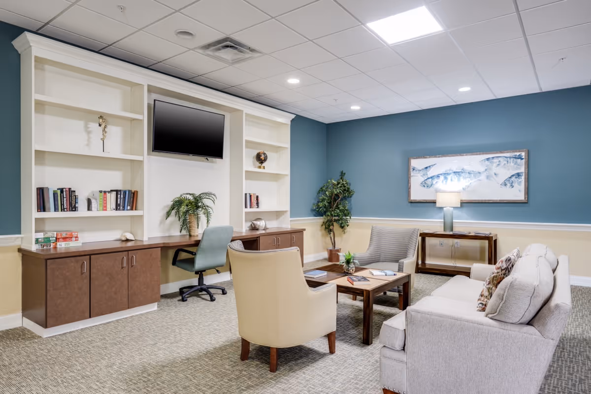 A cozy senior living common area with built-in white shelving and TV, seating around a coffee table, and artwork on blue-green walls.