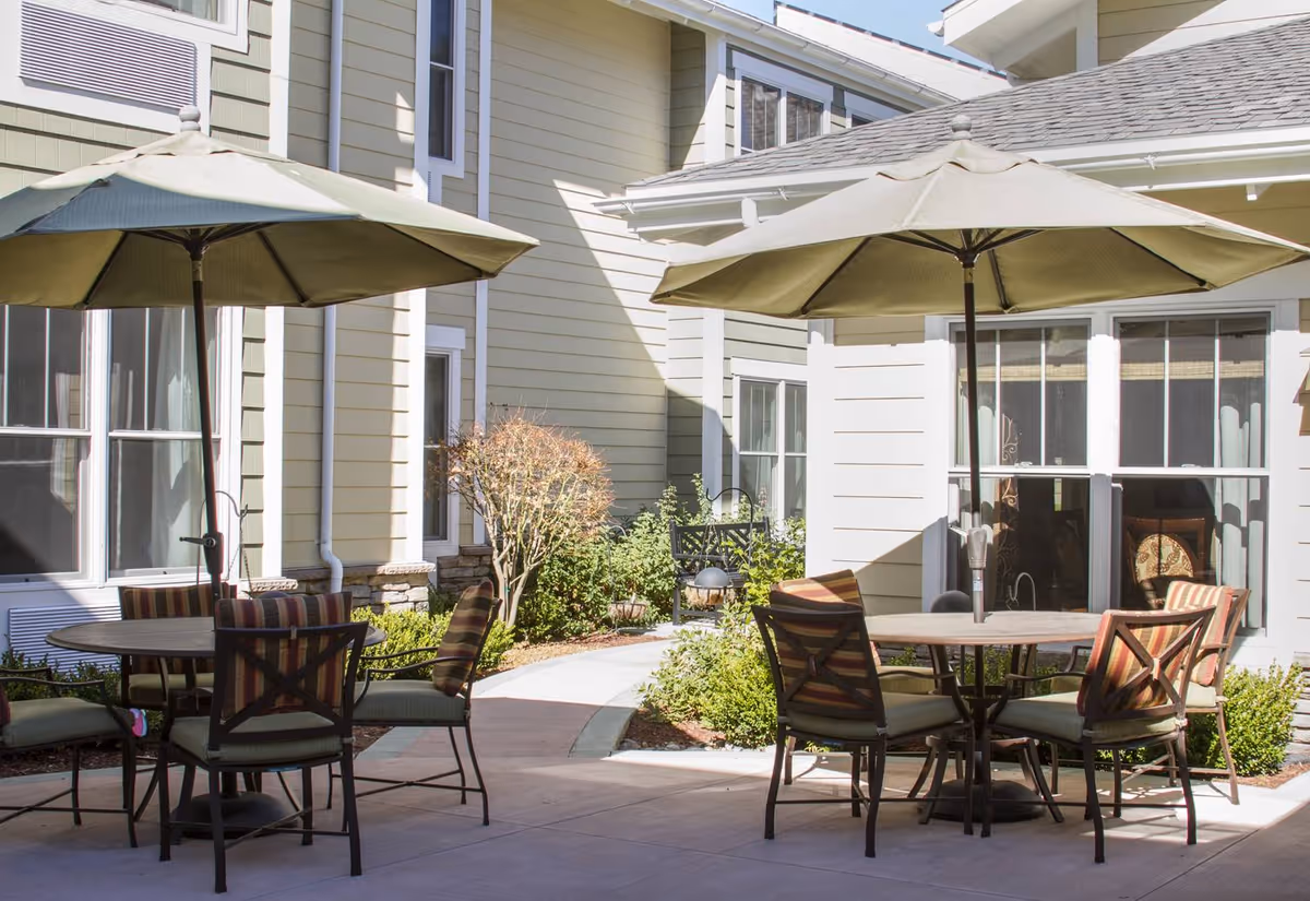 Outdoor patio area at Ivy Park at Monterey with two round tables, each surrounded by four cushioned chairs with striped upholstery. Each table has a large beige umbrella providing shade. The patio is paved and bordered by small bushes and plants, with the beige exterior walls and windows of the building in the background.
