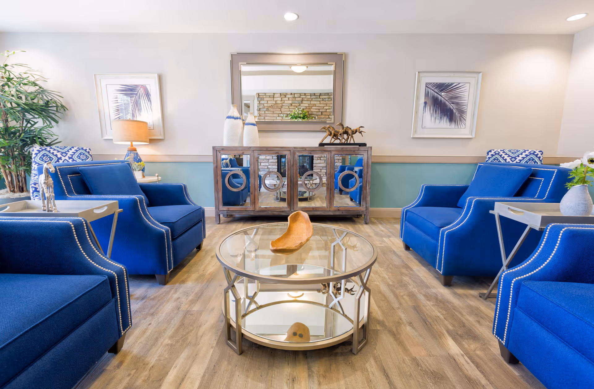 A bright and modern living room with four blue armchairs arranged around a round glass coffee table. The room features wooden flooring, a mirrored cabinet with decorative items, two framed leaf prints on the wall, a table lamp, and a tall green plant in the corner.