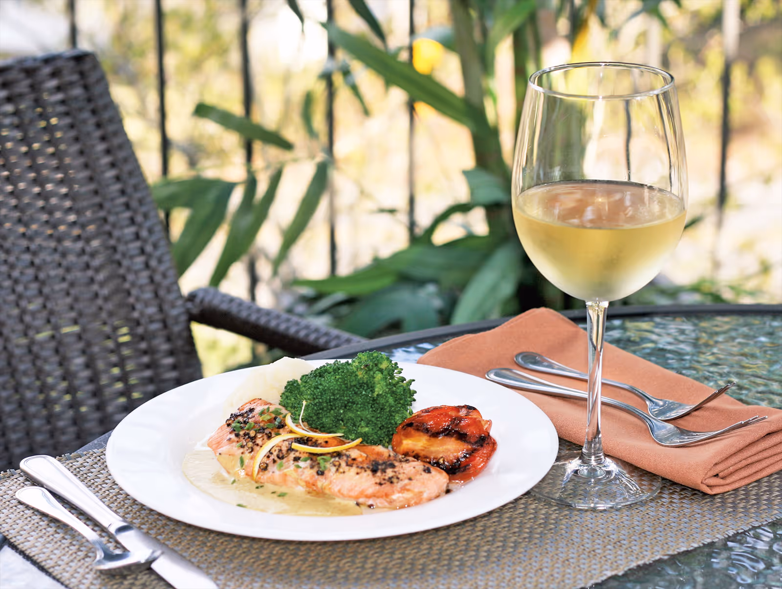 A plate of cooked salmon with broccoli and a grilled tomato slice on a table next to a glass of white wine. The table is set outdoors with a woven chair and green plants in the background.
