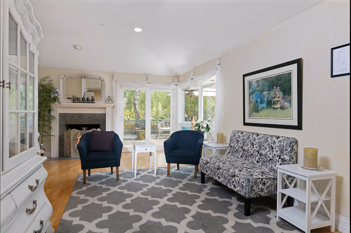 A cozy living room with two dark blue armchairs and a patterned sofa arranged around a white side table on a gray and white patterned rug. There is a fireplace with a decorative screen and a mirror above it on the left side. Large windows and a glass door provide a view of a green outdoor area with trees and patio furniture. The walls are light-colored and decorated with framed artwork. Two white side tables with candles are placed beside the sofa.