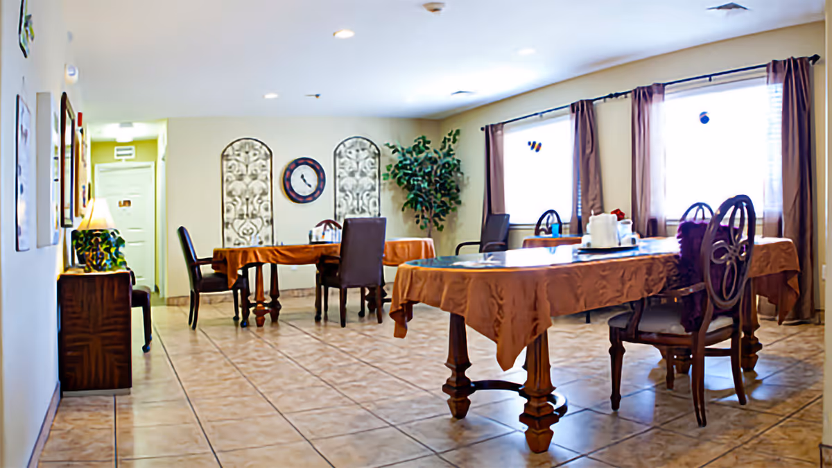 A dining room with two rectangular tables covered with brown tablecloths, surrounded by wooden chairs. The room has tiled flooring, three large windows with brown curtains, a wall clock, decorative wall panels, and a potted plant in the corner. A small wooden cabinet with a lamp is visible on the left side.
