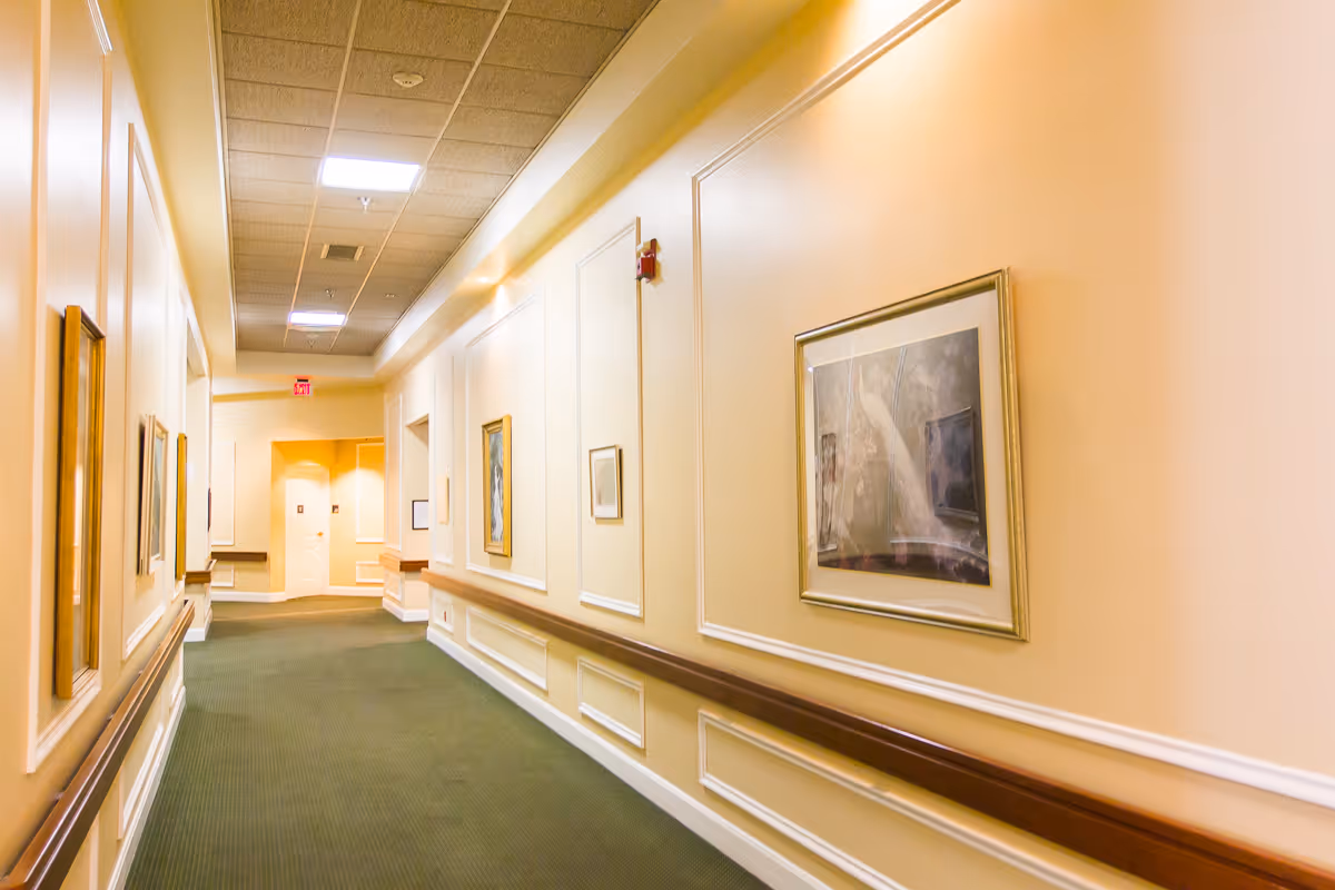 A well-lit indoor hallway with beige walls, green carpet, and framed artwork hanging on the walls. The hallway has a drop ceiling with fluorescent lights and wooden handrails along the walls.