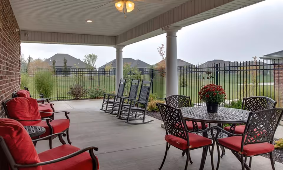 Covered outdoor patio area with red cushioned chairs and a round metal table with a potted plant on top. The patio overlooks a fenced grassy area with houses in the background under a cloudy sky.