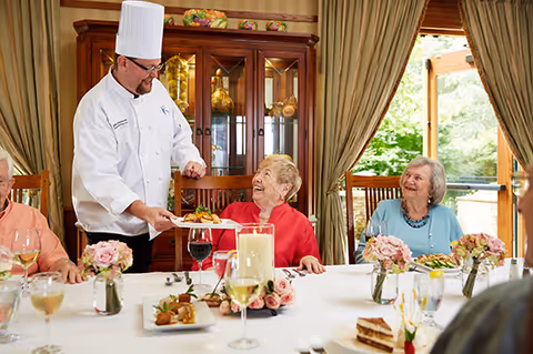 A chef serves a plated meal to smiling elderly residents seated around a decorated dining table in a bright dining room.