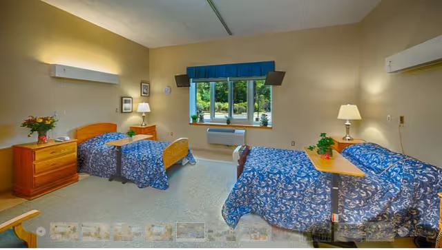 A bright double-occupancy senior care bedroom with two single beds covered in blue patterned bedspreads, bedside tables, lamps, and a window overlooking greenery.