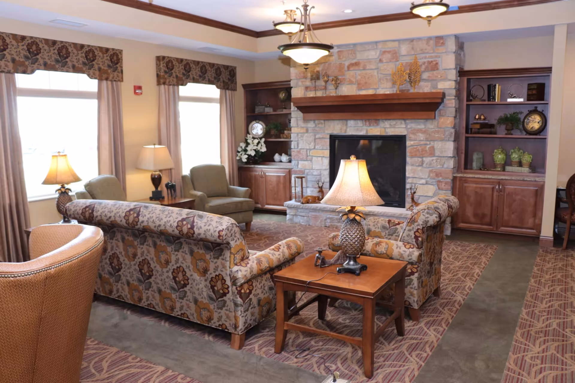 A cozy senior living room with floral patterned sofas, two armchairs, wooden side tables with pineapple-shaped lamps, a stone fireplace, and built-in wooden shelves decorated with clocks, plants, and ornaments. The room has large windows with floral valances and beige curtains, and a patterned carpet covering the floor.