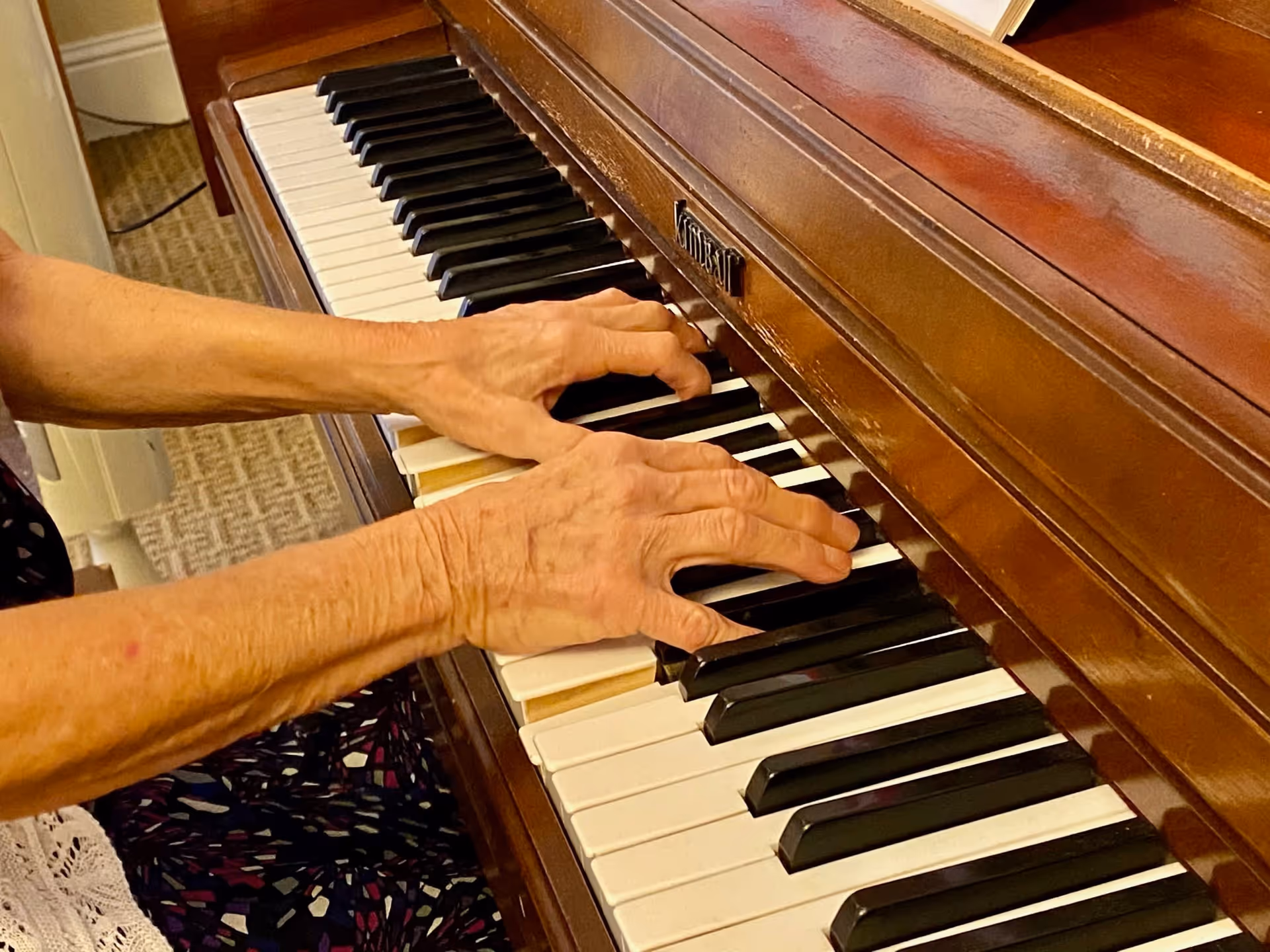 Close-up of an elderly person's hands playing a brown upright piano with visible white and black keys, seated on a patterned carpeted floor.