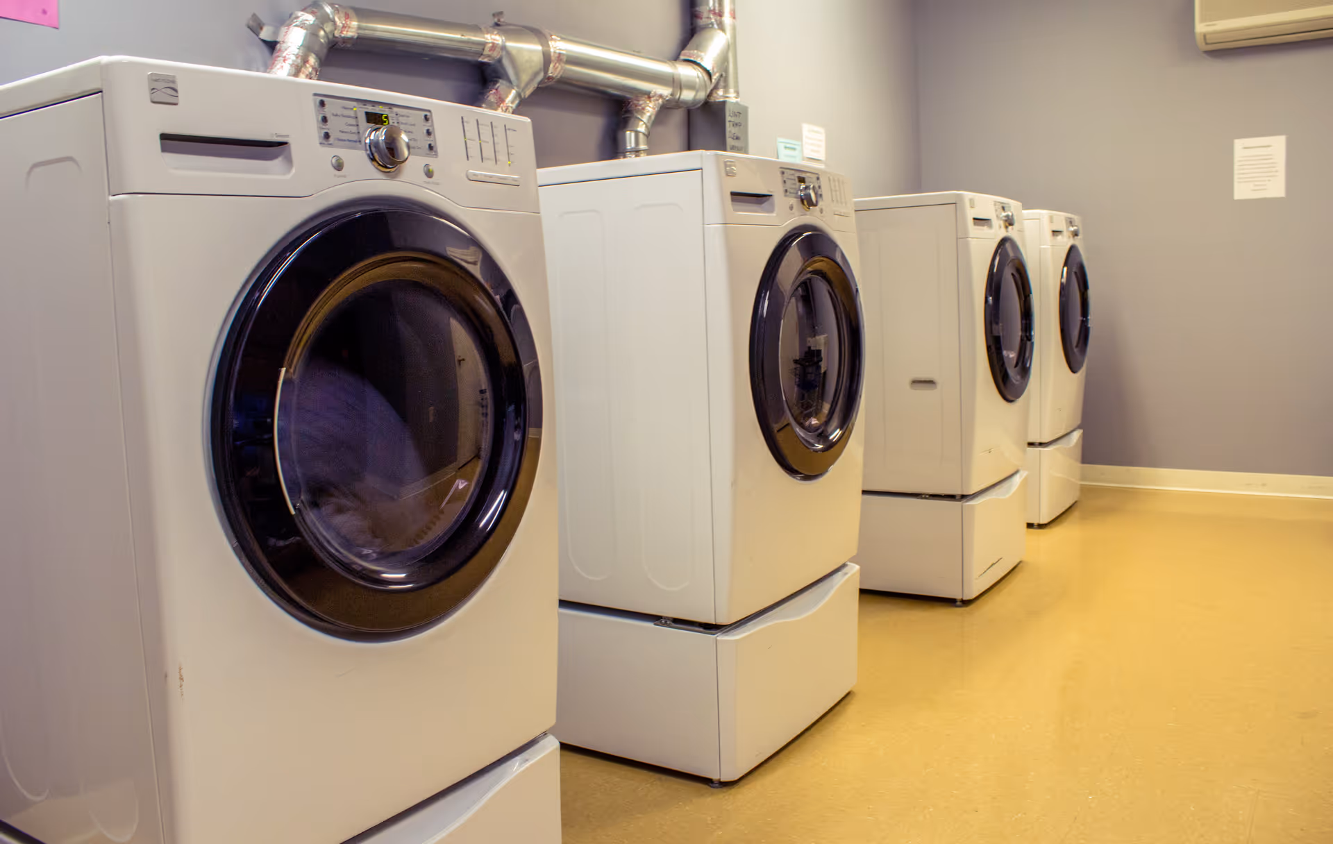 A row of four white front-loading washing machines or dryers in a laundry room with beige flooring and gray walls.