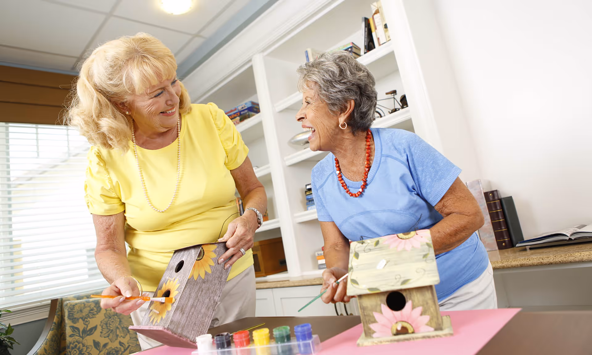 Two elderly women smiling and painting decorative birdhouses at a table in a bright room with shelves and books in the background.