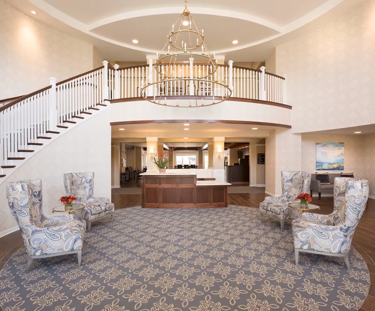 Spacious senior living facility lobby with a large circular patterned carpet, four upholstered armchairs with paisley design arranged around small glass tables with red flower arrangements, a wooden reception desk in the background, a grand staircase with white railings on the left, and a large circular chandelier hanging from the ceiling.