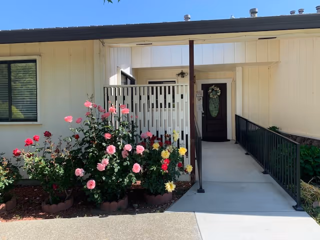 Entrance to a building with a concrete walkway and black metal railing leading to a dark brown door decorated with a floral wreath. To the left of the walkway, there is a garden bed with blooming pink, red, and yellow roses. The building exterior is light-colored with a window on the left side.