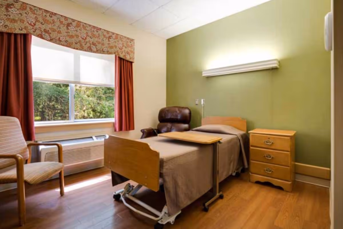 A single room in a senior living facility with a hospital-style bed covered in a brown blanket, a wooden bedside table with three drawers, a brown leather recliner chair, and a wooden armchair near a window with red curtains and a floral valance. The walls are painted light green and cream, and the floor is wooden.