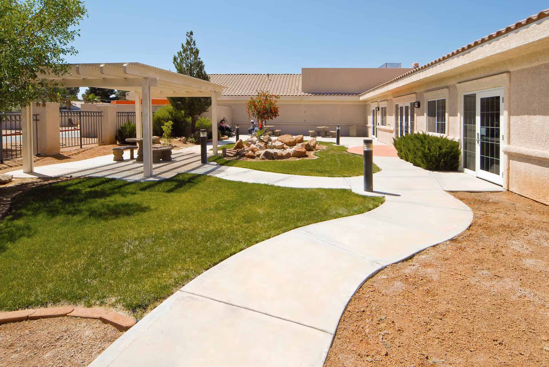 Outdoor courtyard area at Life Care Center of South Las Vegas featuring a curved concrete walkway, green grass, a pergola with benches underneath, a small rock garden with a tree, and the exterior walls and windows of the building under a clear blue sky.