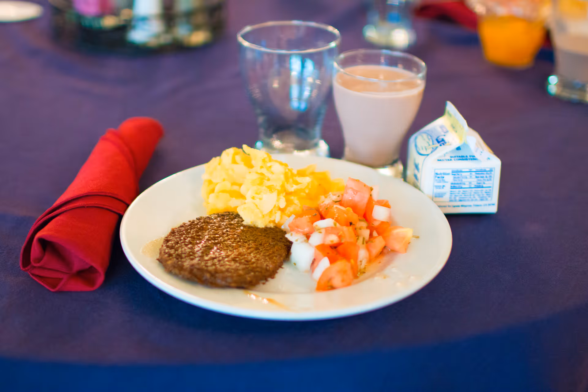 A plate with scrambled eggs, a sausage patty, and a tomato and onion salad on a purple tablecloth. Next to the plate is a red cloth napkin, a glass of chocolate milk, an empty glass, and a small carton of milk.