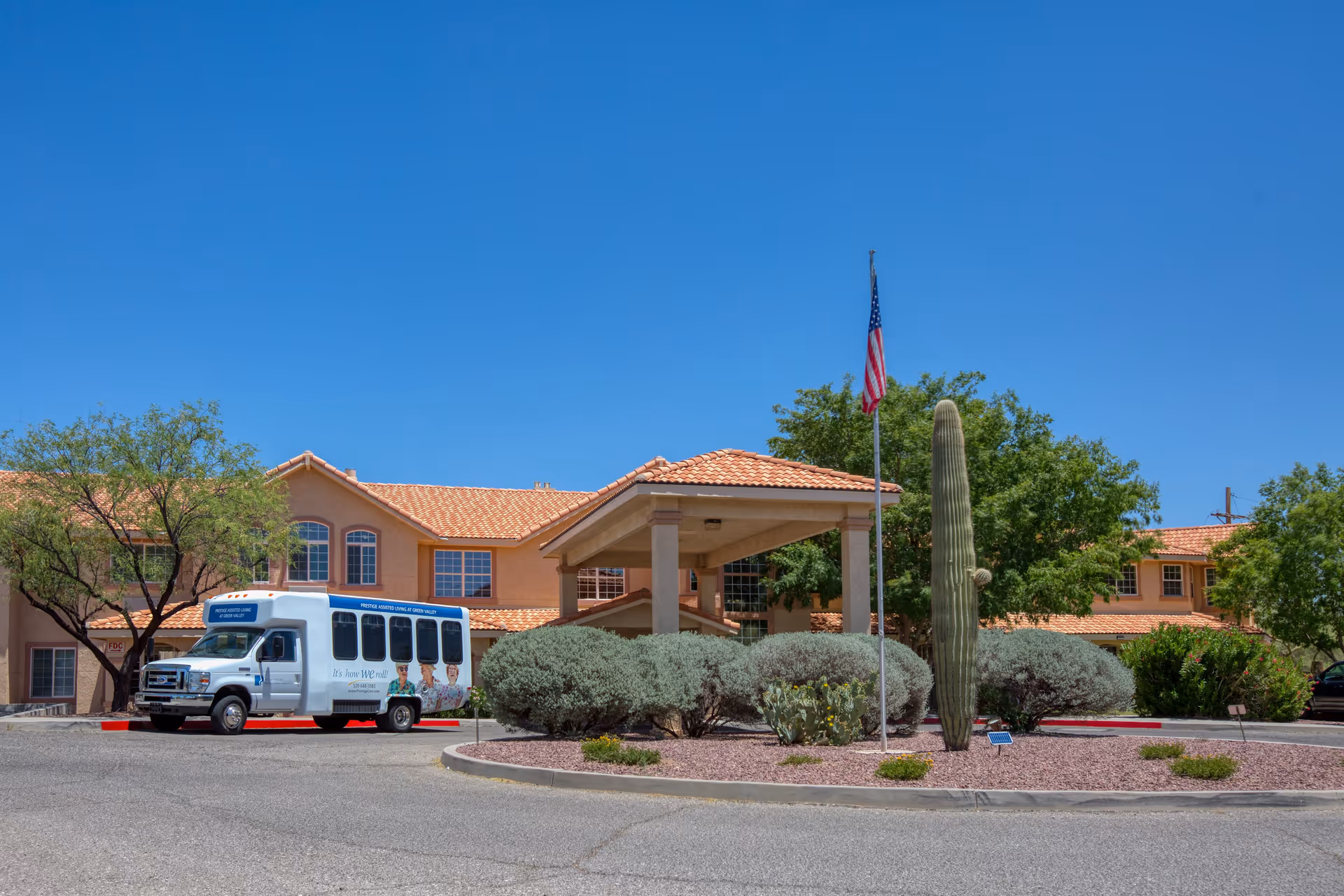Exterior view of Vista Pointe at Green Valley senior living facility with a beige building featuring a tiled roof, an American flag on a flagpole, desert landscaping including a tall cactus, and a white shuttle bus parked near the entrance.