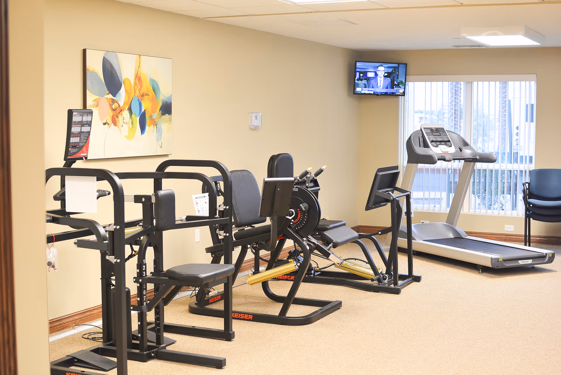 A small fitness room with exercise equipment including a treadmill, a rowing machine, and other workout machines. There is a wall-mounted TV in the corner and a large window with vertical blinds letting in natural light. A colorful abstract painting is hung on the beige wall.