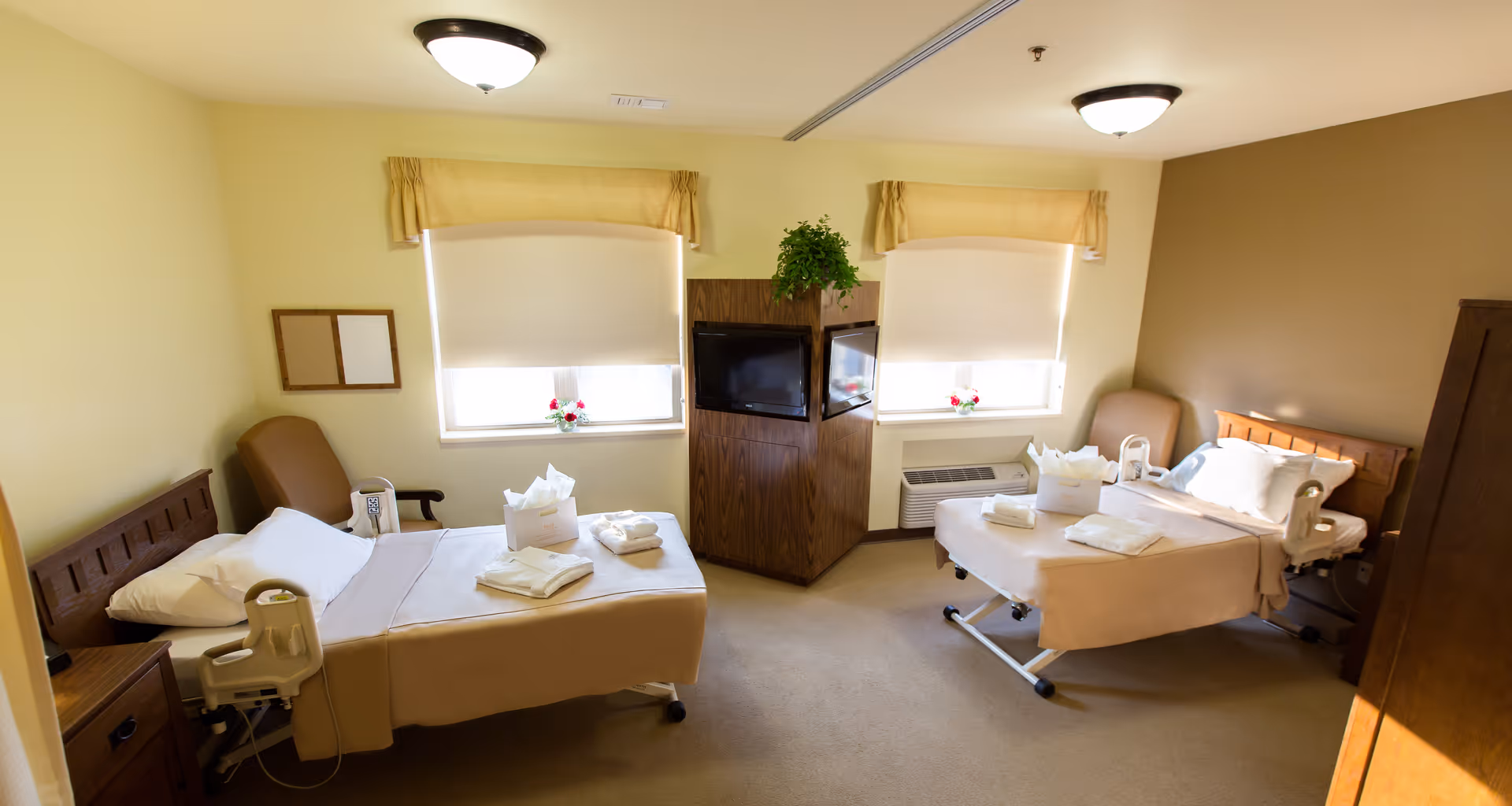A bright, clean room with two hospital-style beds, each with beige bedding and white pillows. Each bed has folded towels and a tissue box placed on top. Between the beds is a wooden cabinet with a TV mounted on it and a green plant on top. Two windows with beige valances and white blinds let in natural light. There are two beige armchairs, one next to each bed, and the walls are painted in light yellow and beige tones.