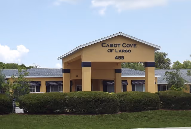 Exterior front view of Cabot Cove of Largo building with a yellow facade, a covered entrance supported by columns, and the address number 455 displayed. The building is surrounded by green bushes and trees under a partly cloudy sky.