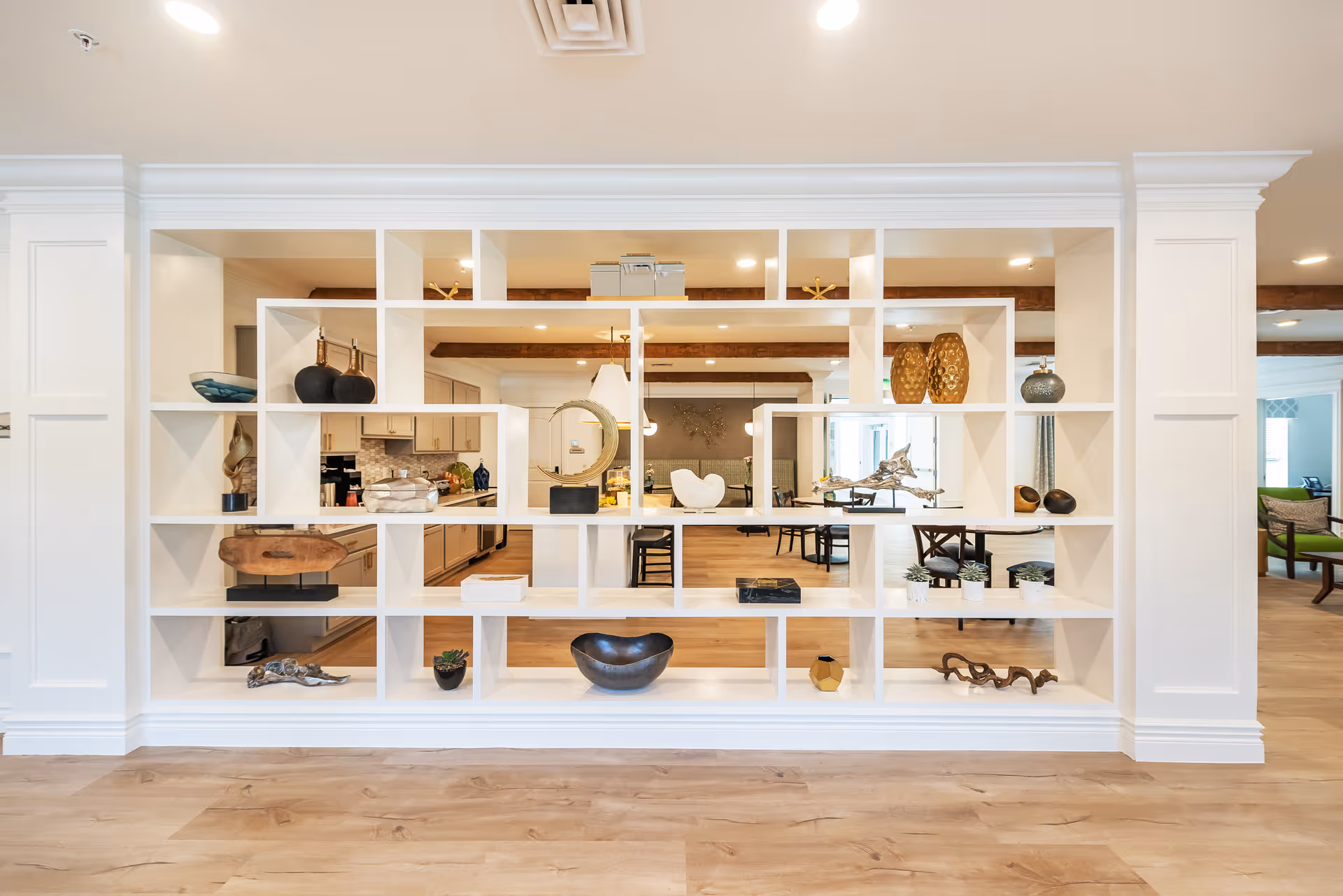 A modern interior space featuring a large white open shelving unit with various decorative items such as vases, bowls, and sculptures. The shelving unit partially divides the room, revealing a kitchen area with cabinets and a dining area with tables and chairs in the background. The floor is light wood, and the ceiling has recessed lighting and exposed wooden beams.