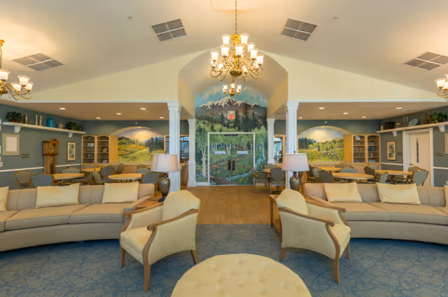 A spacious and well-lit common area in a senior living facility featuring comfortable beige sofas and armchairs arranged around a round ottoman. The room has blue carpeting, wooden flooring in the center, and walls painted with scenic murals of mountains and countryside. There are chandeliers hanging from the ceiling, table lamps on side tables, and built-in shelving units along the walls.