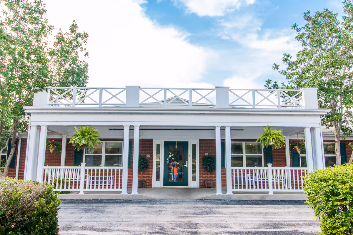Front exterior view of a single-story brick building with white columns and railings, green door with a wreath, hanging plants, and trees on either side under a partly cloudy sky.