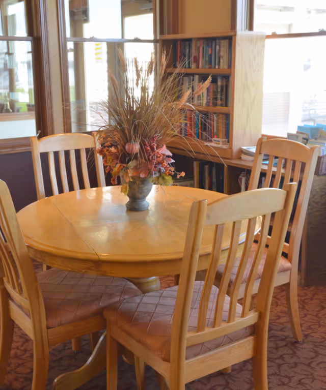 A round wooden table with a decorative floral arrangement in a vase at the center, surrounded by four wooden chairs with cushioned seats. In the background, there is a bookshelf filled with books and large windows letting in natural light.
