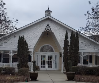 Front exterior view of a senior living facility building with a peaked roof, white siding, and decorative trim. There are tall evergreen shrubs and potted plants flanking the entrance, and leafless trees on either side under a cloudy sky.