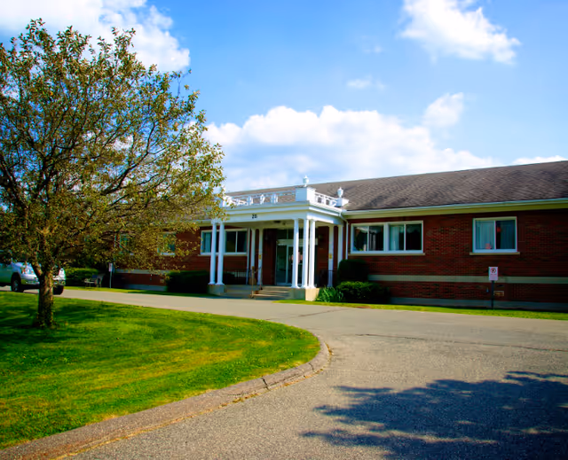 Single-story brick nursing facility with a white columned entrance, curved driveway and lawn under a blue sky.