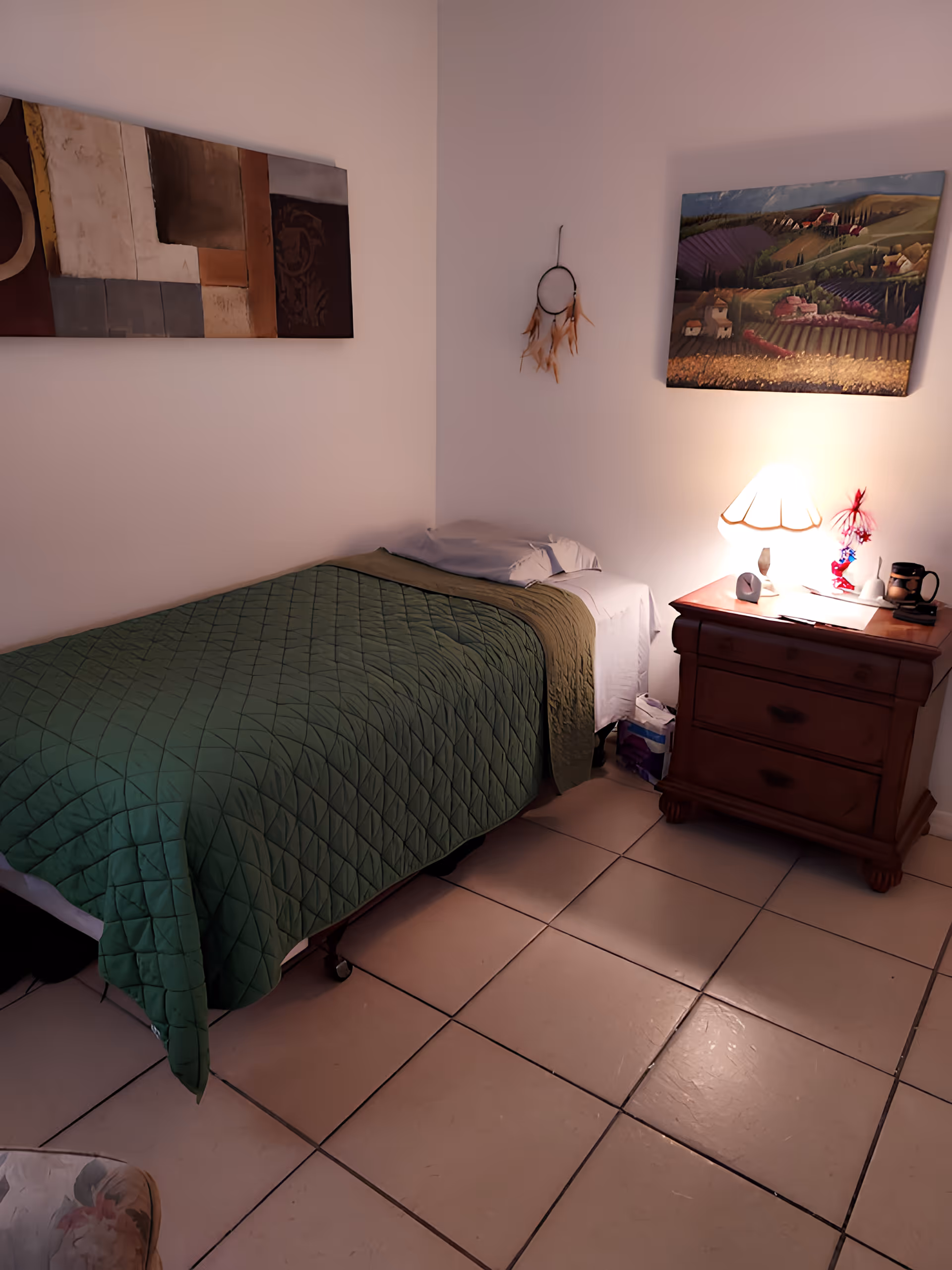 A small bedroom with a single bed covered with a green quilt. Next to the bed is a wooden nightstand with a lit lamp, a mug, and some small items. The walls are decorated with two paintings and a hanging dreamcatcher. The floor is tiled.