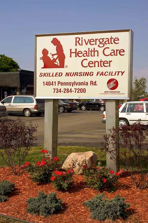 A large Rivergate Health Care Center sign with landscaping in a parking lot at the facility.