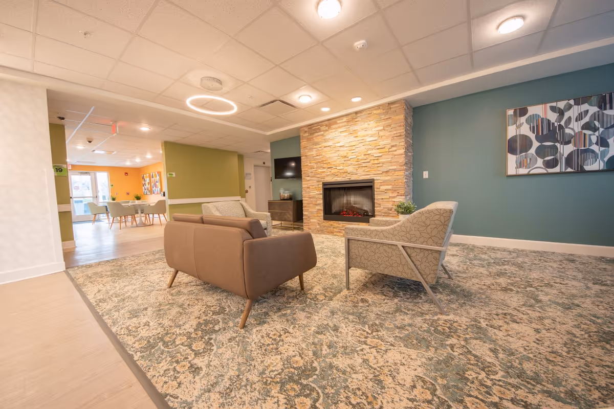 A cozy senior living common area with a patterned carpet, two armchairs, and a brown sofa arranged around a modern stone fireplace. The walls are painted teal and green, with a colorful abstract painting hanging on one wall. In the background, there is a dining area with tables and chairs near large windows letting in natural light.