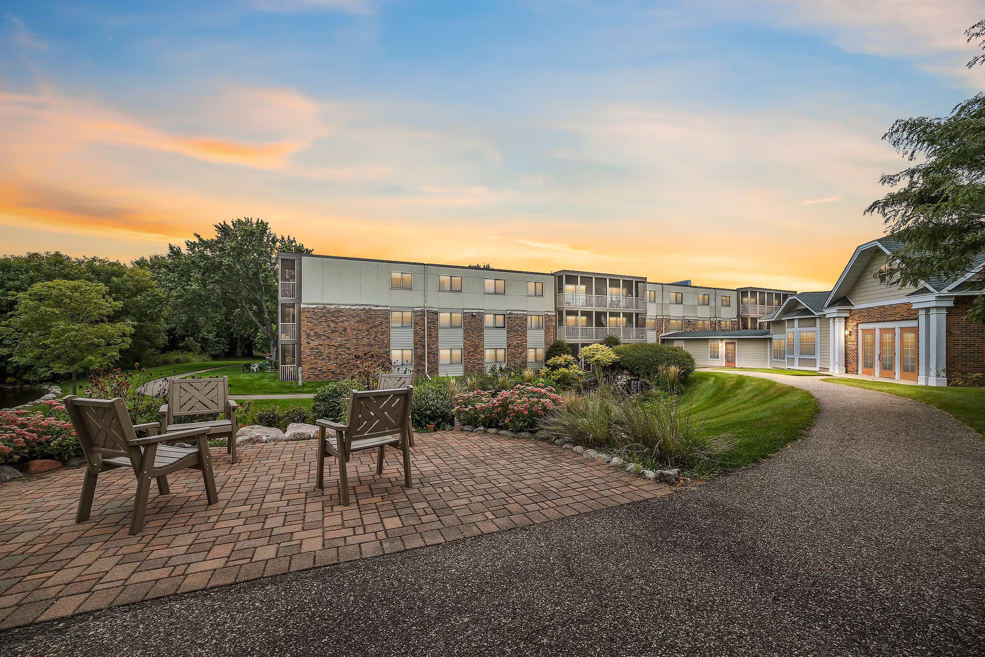 Outdoor patio area with four wooden chairs arranged on a brick-paved surface, surrounded by greenery and flowers. In the background, there is a multi-story building with balconies and large windows, and a pathway leading to another building with large glass doors. The sky is colorful with a sunset glow.