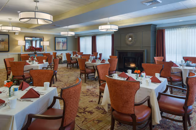 A spacious dining room with multiple tables covered in white tablecloths, each set with folded red napkins, cups, and small flower arrangements. The room features comfortable upholstered chairs, large windows with sheer curtains, a fireplace, and modern ceiling lights.