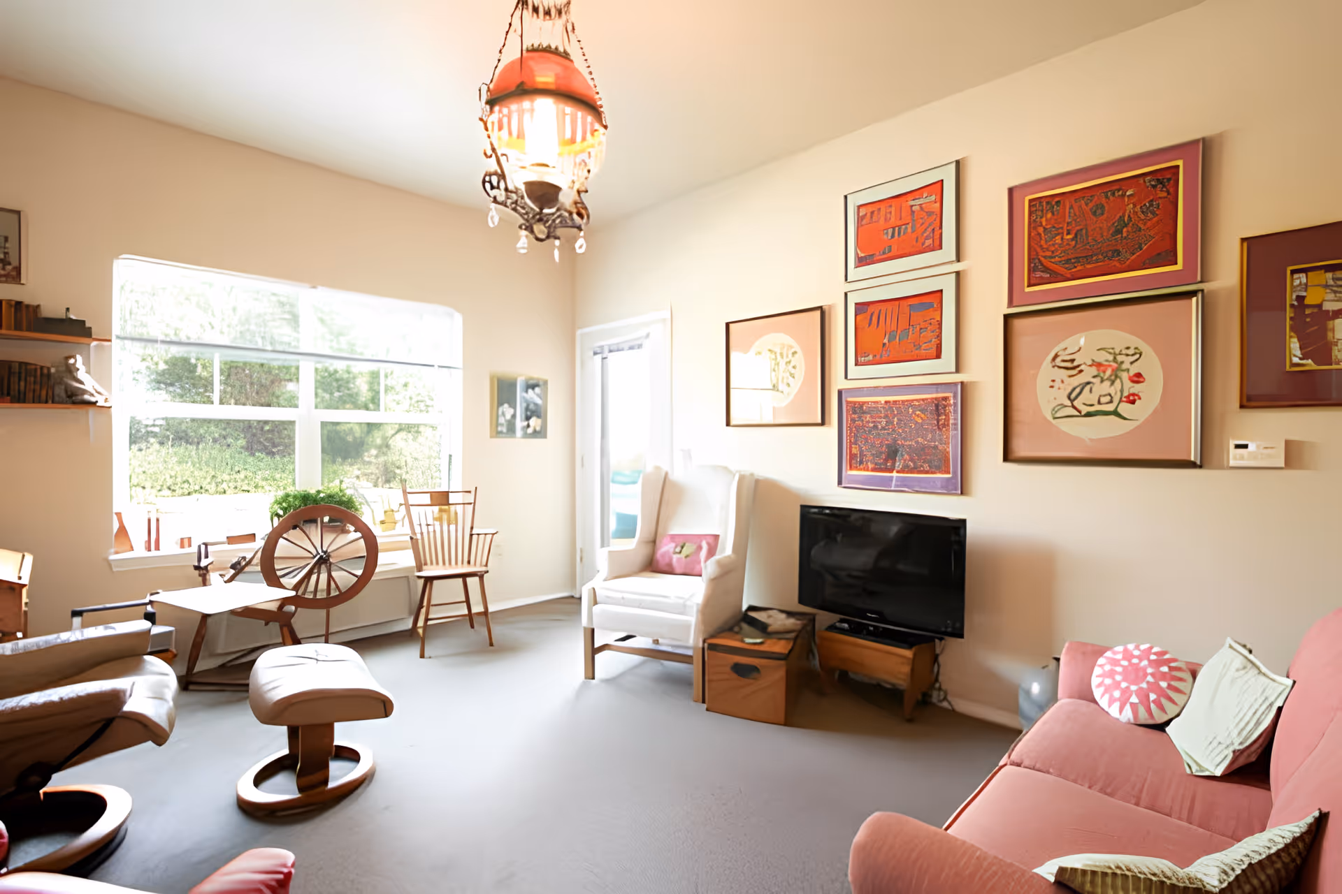 A bright living room with a large window letting in natural light. The room features a pink sofa with decorative pillows, a white armchair, a wooden rocking chair, and a small TV on a wooden stand. The walls are adorned with colorful framed artwork, and a vintage-style hanging lamp is suspended from the ceiling. There is also a wooden spinning wheel near the window.