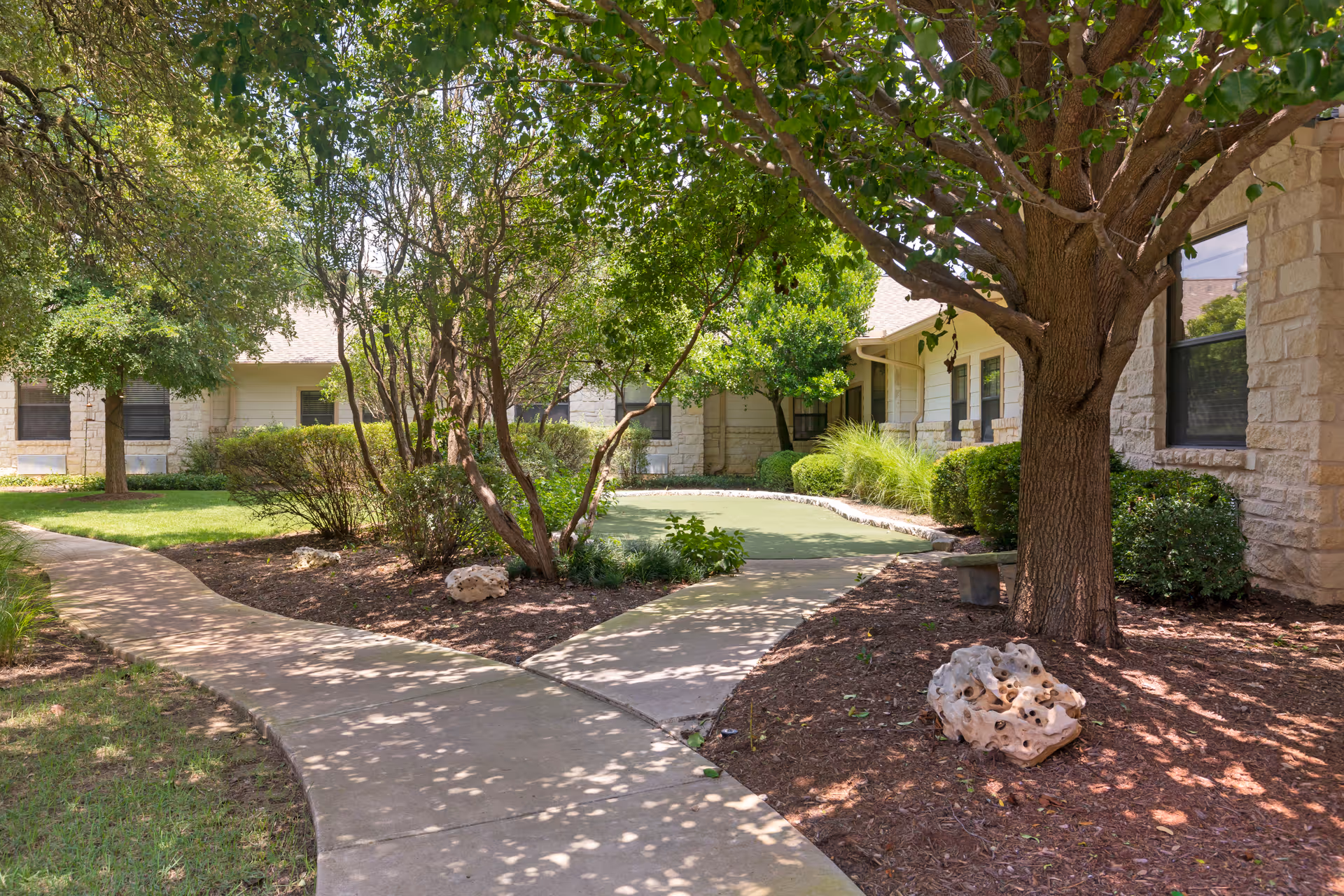 A shaded outdoor courtyard area with a concrete pathway winding through green grass, trees, and bushes. The courtyard is surrounded by a light-colored stone building with several windows. There is a large tree casting shadows on the ground and a rock placed near the pathway.