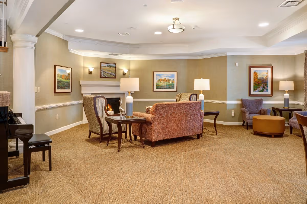Comfortable communal living room with upholstered chairs and sofas arranged around a fireplace, lamps, framed artwork, and a piano.