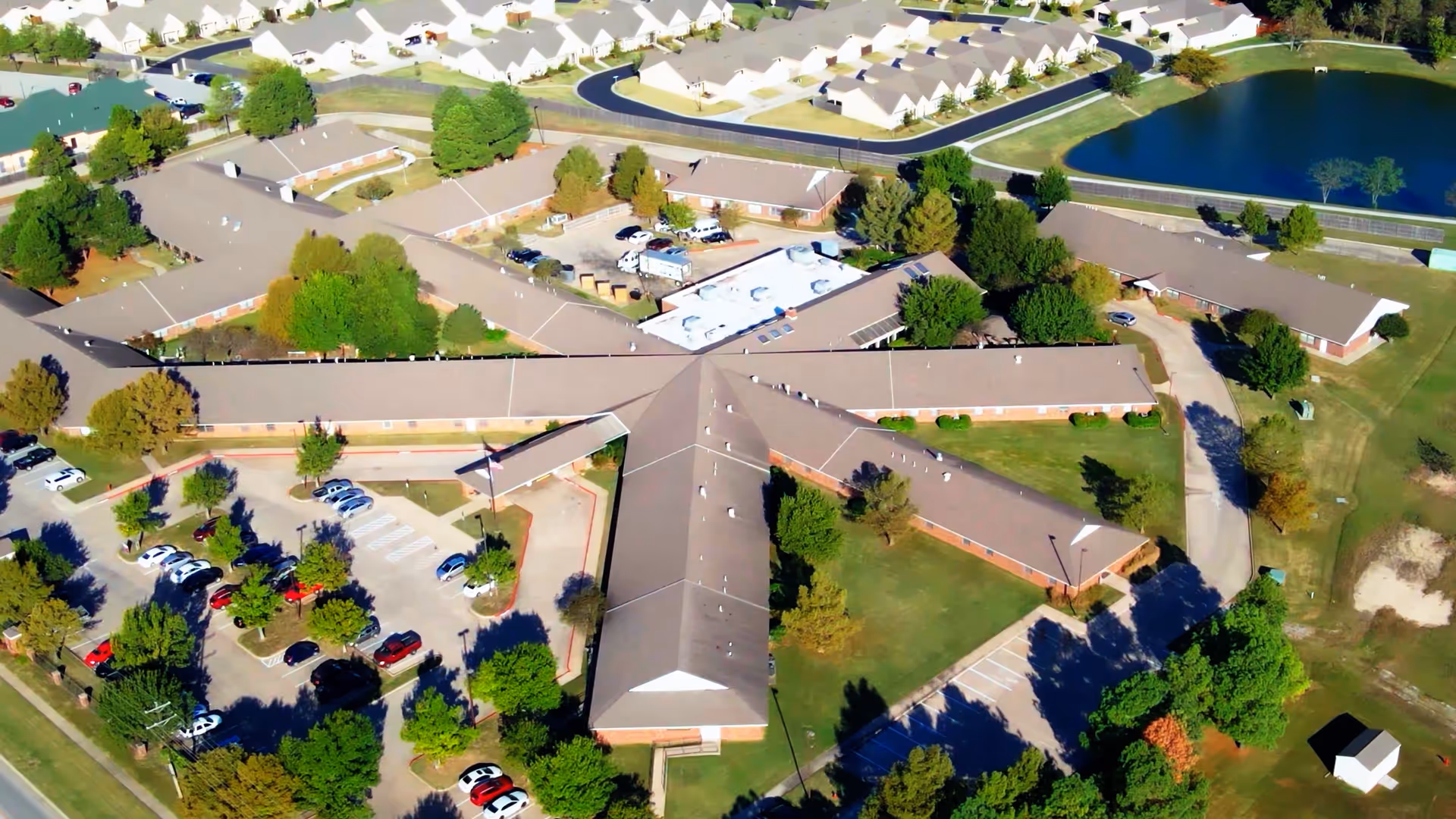 Aerial view of a single-story assisted living complex with connected buildings, parking lots, trees, and a nearby pond.