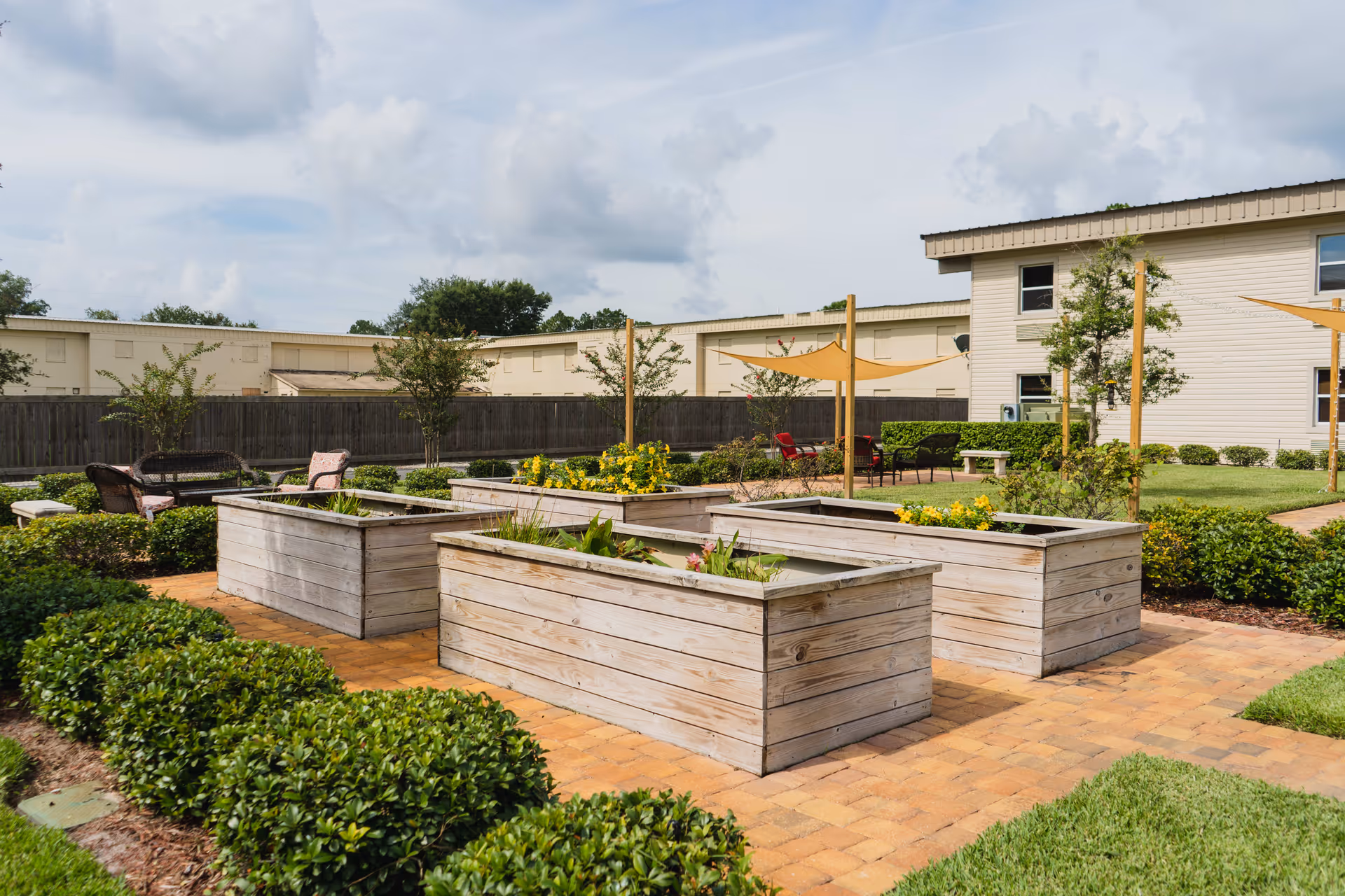 Outdoor garden area with raised wooden planter boxes containing various plants and flowers, surrounded by neatly trimmed bushes and paved walkways. In the background, there are seating areas with chairs and shade sails, and a light-colored building with windows.