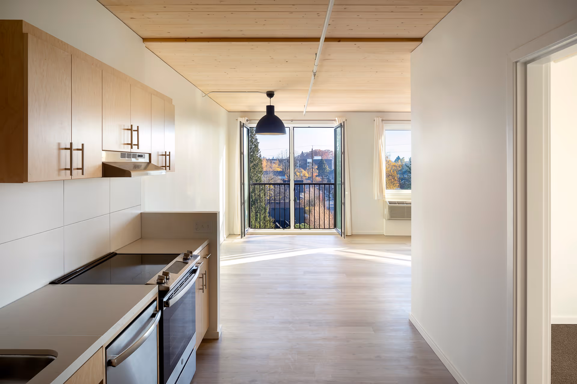 A bright, modern kitchen area with light wood cabinets, a stainless steel oven and dishwasher, and a light countertop. The kitchen opens into a spacious living area with large windows and a glass door leading to a balcony with a black railing. The ceiling is wooden with a black pendant light hanging in the living area. Outside, trees and buildings are visible under a clear sky.