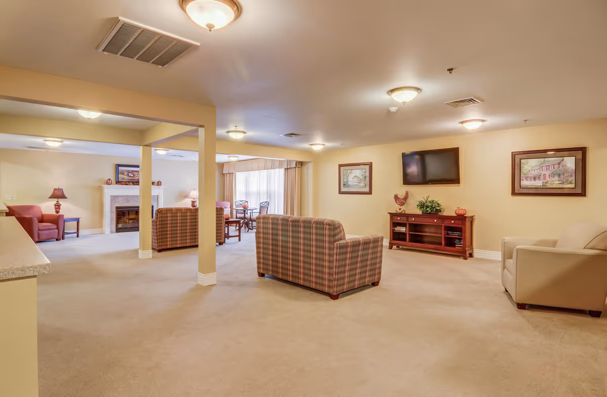 A spacious living room area in a senior living facility with beige walls and carpet. The room features multiple seating options including plaid armchairs, a beige armchair, and a red armchair. There is a wooden TV stand with a flat-screen TV mounted above it, two framed paintings on the wall, and a fireplace in the background. The room is well-lit with ceiling lights and natural light coming through large windows with curtains.