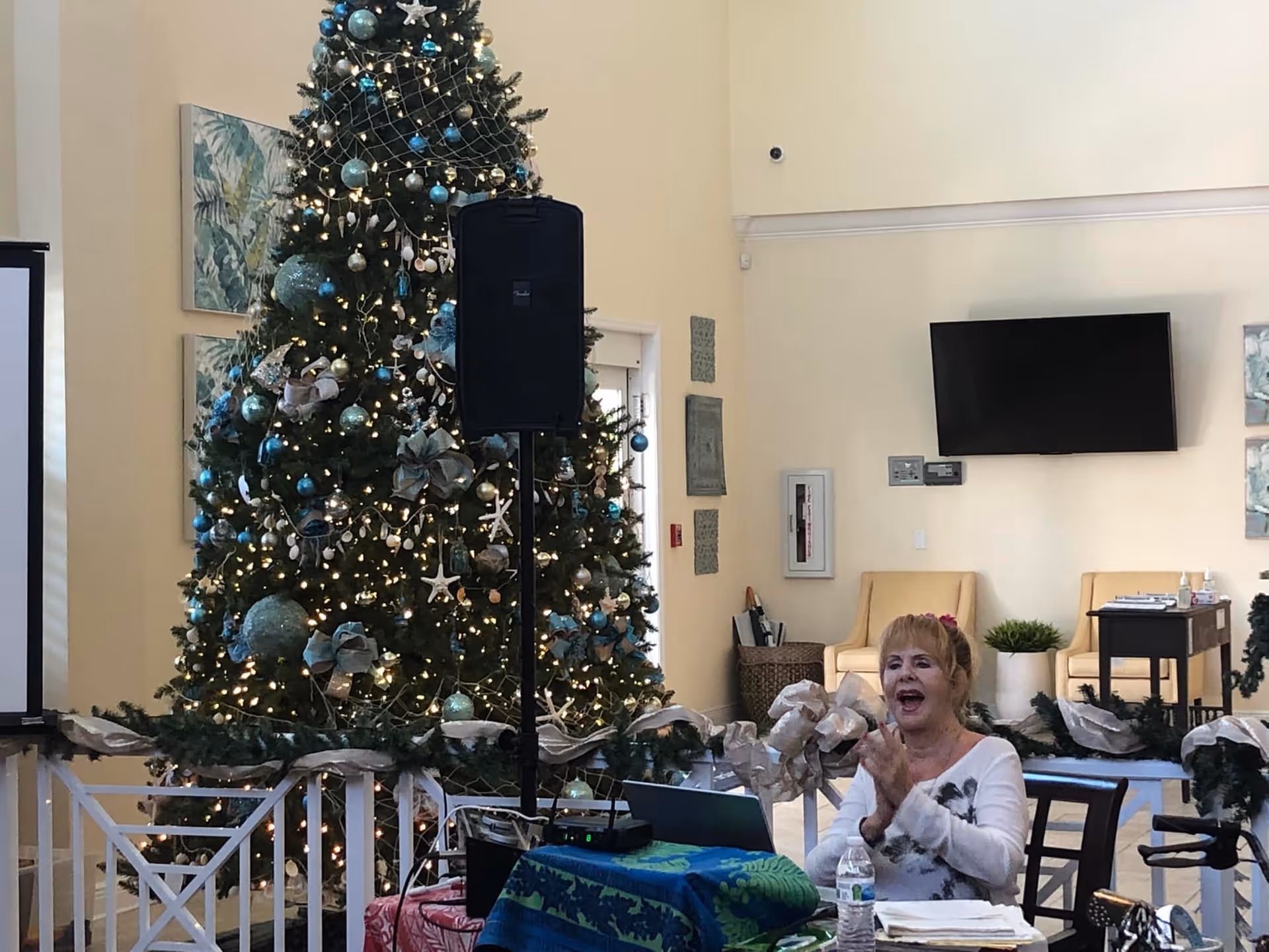 A decorated Christmas tree with blue and silver ornaments and lights stands in a room with beige walls. In front of the tree, a woman with blonde hair is sitting at a table covered with a blue and green cloth, clapping her hands. The room has chairs, a wall-mounted TV, and some wall decorations.