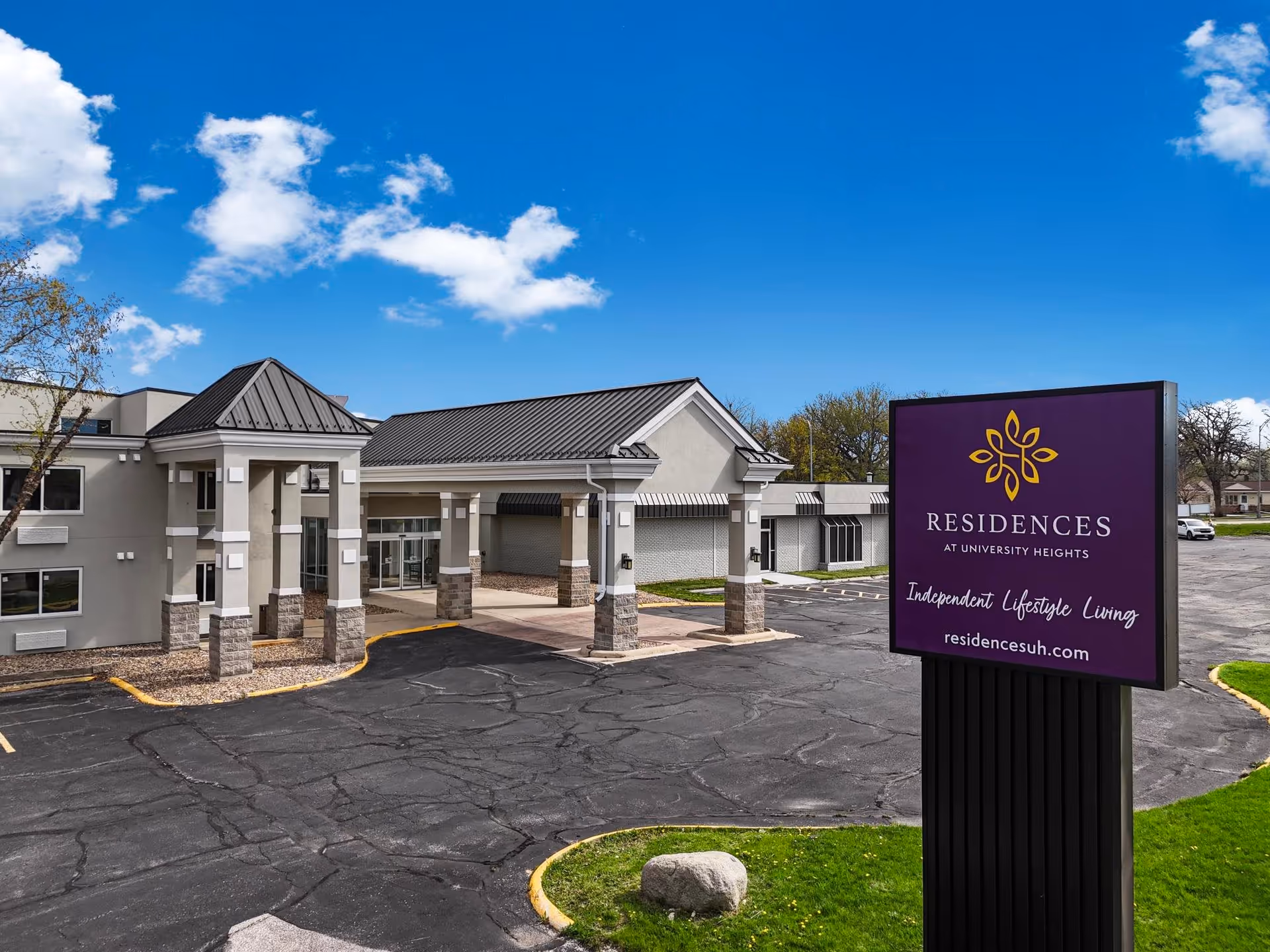 Exterior view of the Residence at University Heights building with a covered entrance and a large purple sign in the foreground displaying the facility name, logo, and website under a blue sky with some clouds.