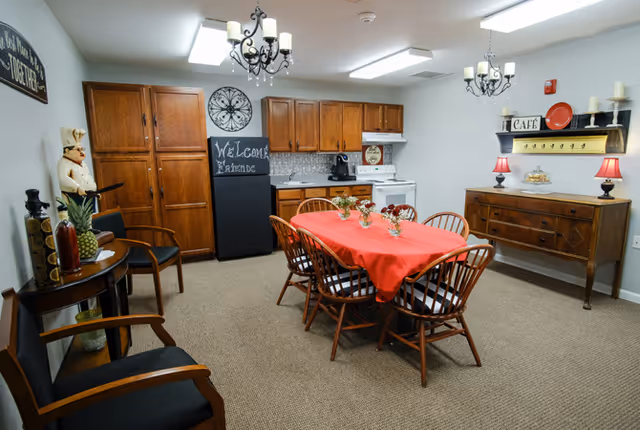 A cozy dining area in a senior living facility with a wooden table covered by a red tablecloth and six wooden chairs. The room features wooden cabinets, a small kitchen area with a stove and coffee maker, decorative wall art including a chalkboard with 'Welcome Friends' written on it, and a sideboard with lamps and decorative items. The space is well-lit with ceiling lights and chandeliers.